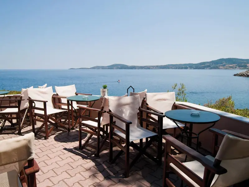 Wooden directors' chairs with cream canvas backs and small green tables on a paved terrace, the gulf and far hills across the water