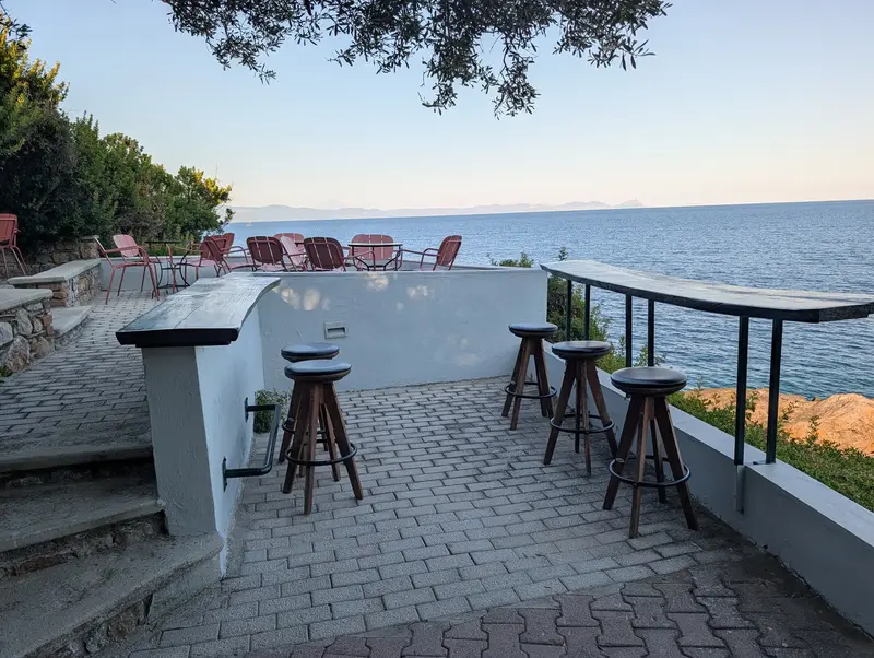 The bar counter with wooden stools and a low white wall, the sea going grey-blue at dusk, olive branches overhead and red metal chairs on the terrace beyond