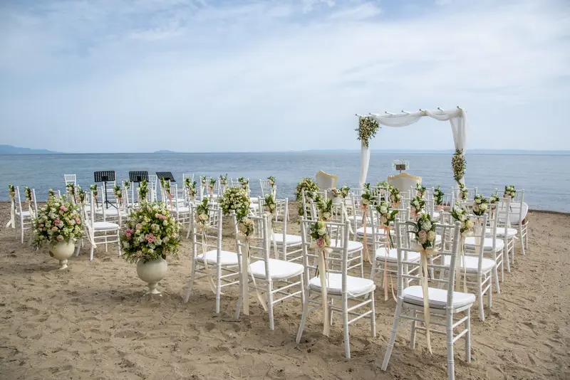 Rows of white chairs on the sand facing a floral arch at the water's edge, the gulf and a far headland behind it