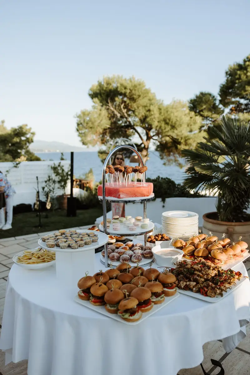 A white-clothed table of canapés on tiered stands, set out in the open air with pines and the water behind