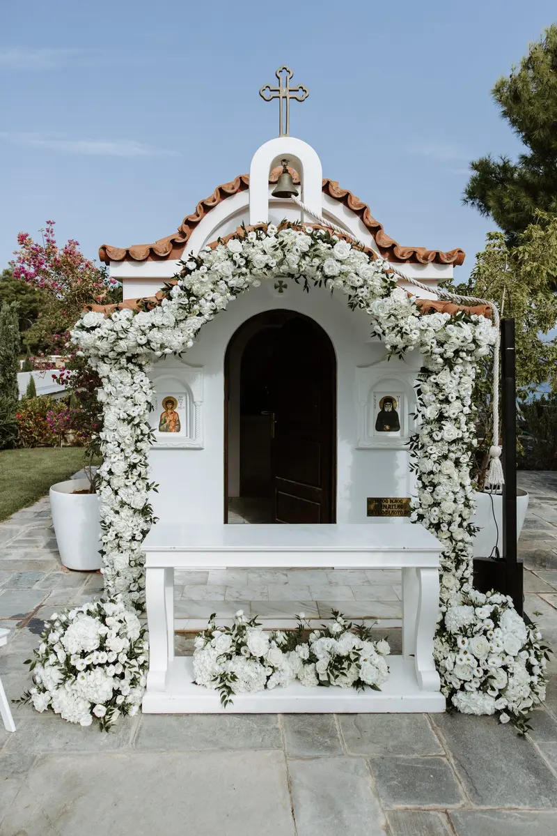 A small whitewashed chapel with a tiled roof, its doorway framed by an arch of white roses and greenery, under a clear blue sky