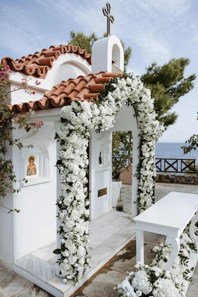 The chapel doorway from the side, white flowers running up the arch and around the bell, the sea visible past the corner of the building