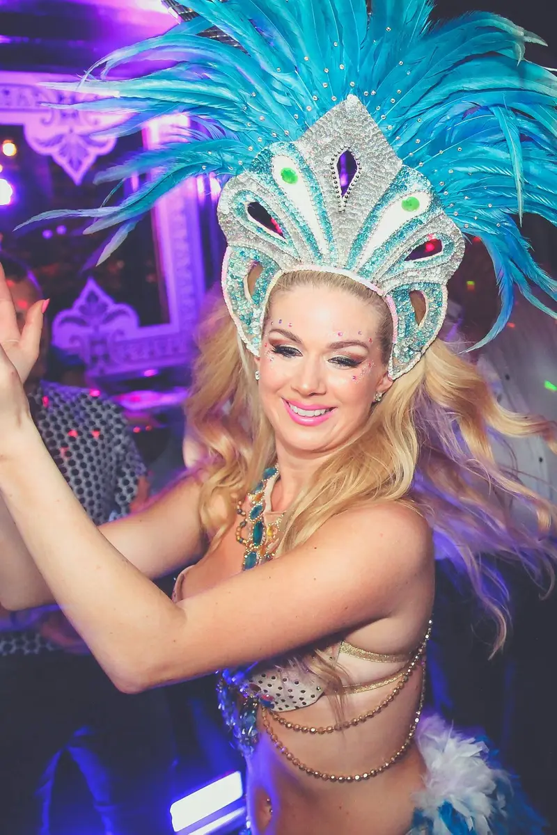 A carnival dancer in a turquoise feather headdress, close, under violet light