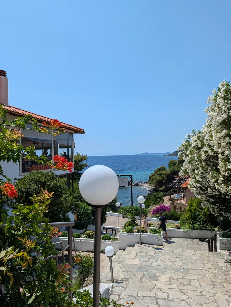 Steps down through oleander to a taverna terrace above a small cove, the Aegean and a headland beyond