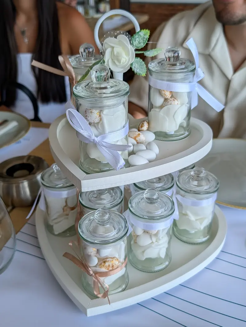 A tiered heart-shaped stand of glass jars filled with white sugared almonds, tied with white and blush ribbons, a single white rose above them