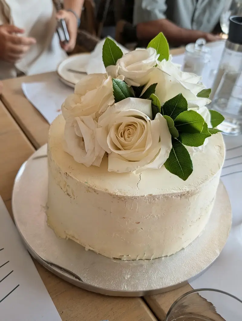 A single-tier white cake, roughly finished in buttercream and topped with cream roses and glossy green leaves