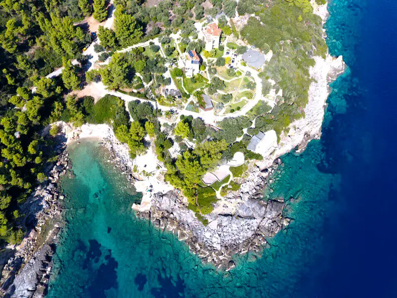 The property from the air, a pine-covered headland with paths and terraces threaded through the trees, a small cove on one side and deep blue water on the other