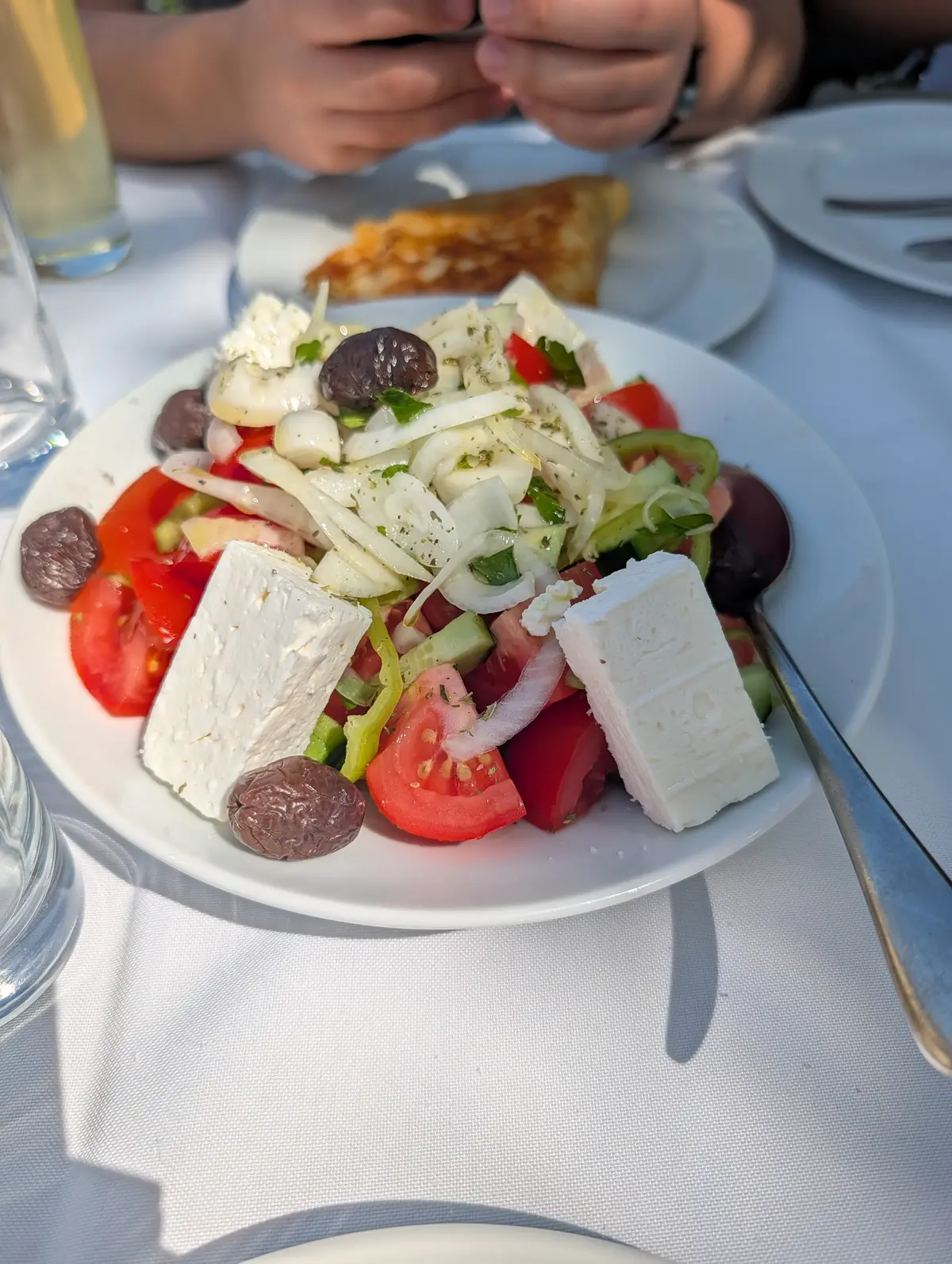 A Greek salad of tomato, cucumber, onion and olives under two thick slabs of feta, a wedge of pie on a plate behind it