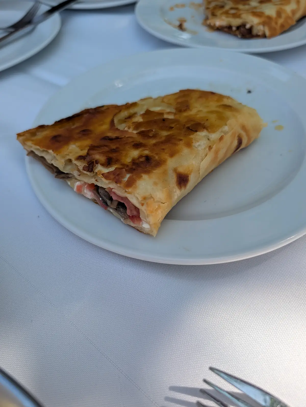 A wedge of Pontian savoury pie on a white plate, its pastry blistered and golden, filled with mushrooms, tomato and soft cheese