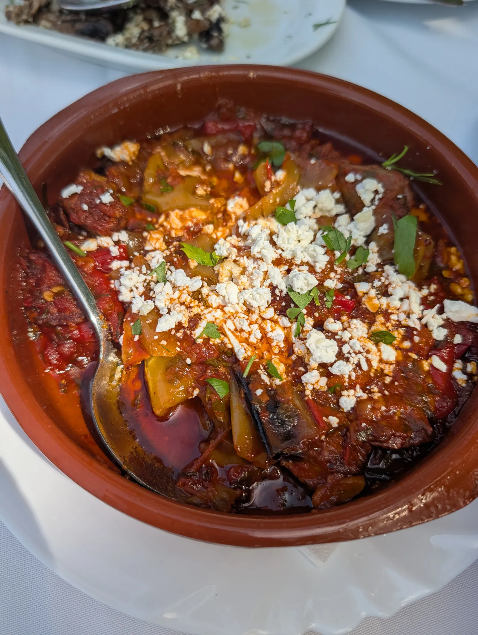 Baked aubergine and peppers in tomato sauce in a terracotta dish, crumbled feta and parsley scattered over the top