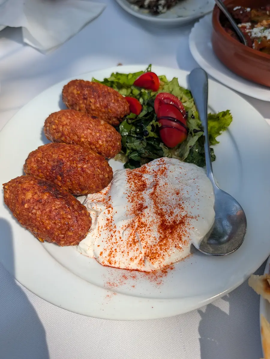 Four fried bulgur kofta beside a spoonful of yoghurt dusted with paprika, with a green salad and tomato