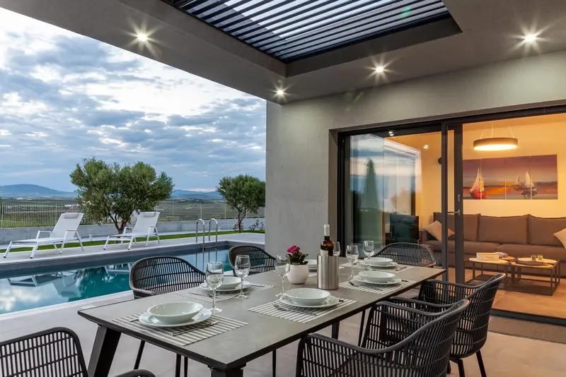 A covered terrace laid for dinner under a slatted pergola, the pool and olive trees beyond, hills fading into evening