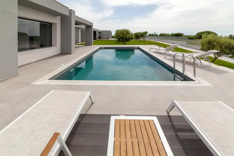 The pool in full daylight, pale stone terrace, sun loungers along one side and olive trees at the far end