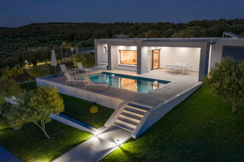The villa at dusk from above, warm light spilling from the open living room and the lit pool reflecting it