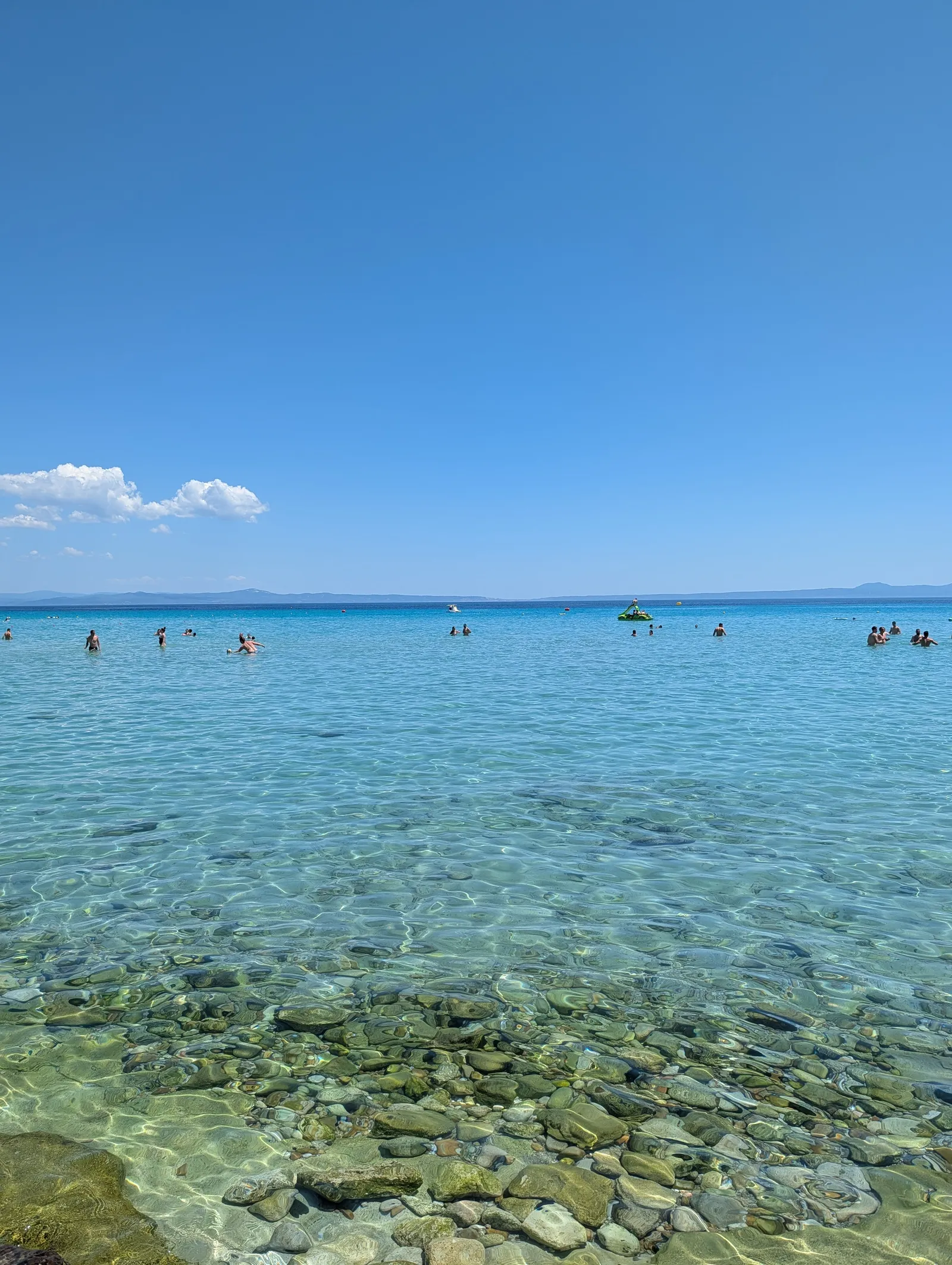Clear turquoise water over pale pebbles on a Kassandra beach, swimmers offshore and the far coastline low on the horizon
