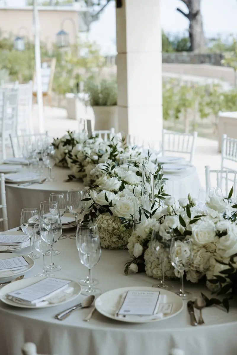 A round table set with white linen and glassware, a tall arrangement of white roses and olive foliage at its centre