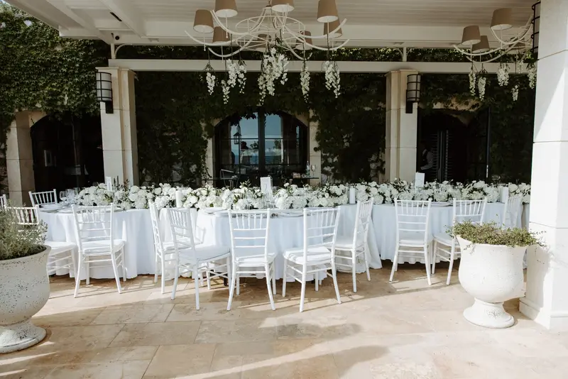 A colonnaded terrace laid for dinner, white chiavari chairs at long tables, the sea flat and pale beyond the columns