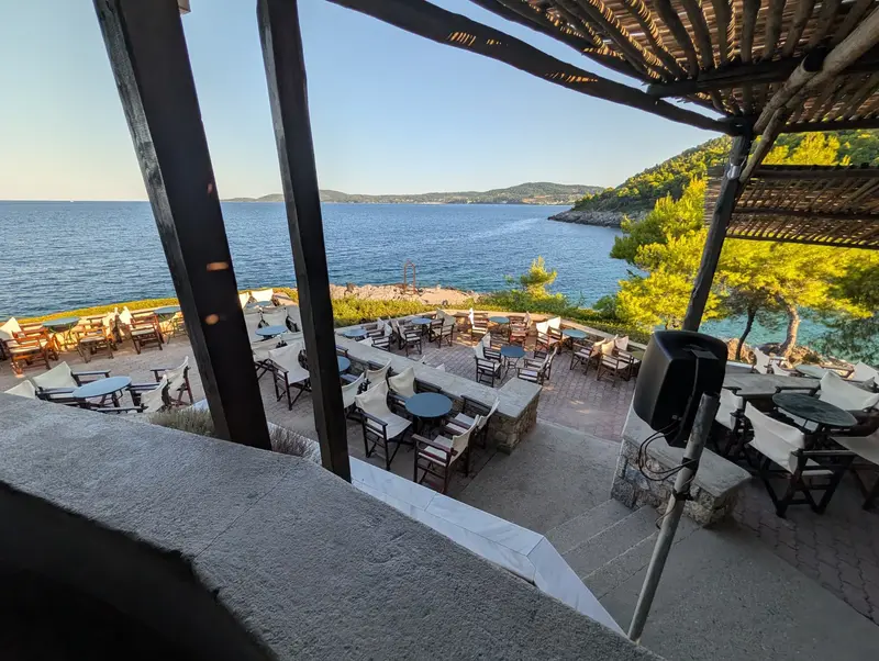 The cliffside terrace under a slatted pergola at golden hour, round tables and dark wood chairs with cream canvas backs, the cove and a green headland beyond