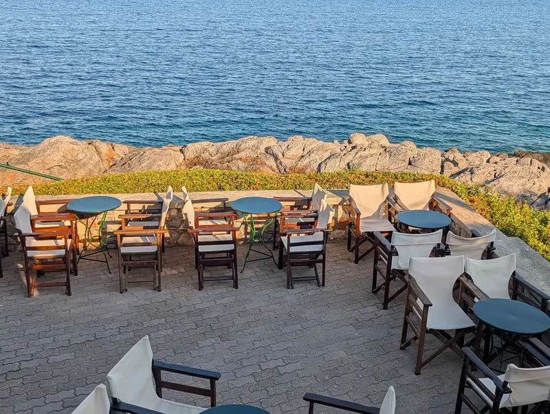 Round tables and canvas-backed wooden chairs on a paved terrace behind a low stone wall, rocks and flat blue sea directly below