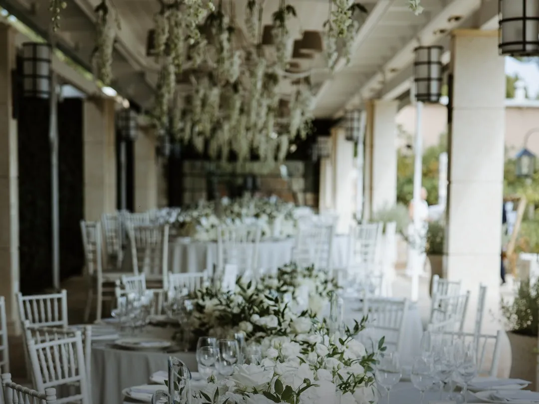 A long table set for a wedding on a colonnaded terrace, white roses down its length and white blossom hanging from the ceiling above