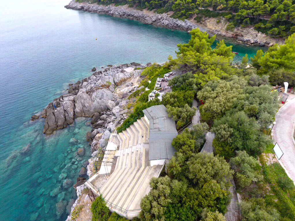 Looking down over the stone steps of the amphitheatre to boulders and clear turquoise shallows, olive trees and a path on the slope above