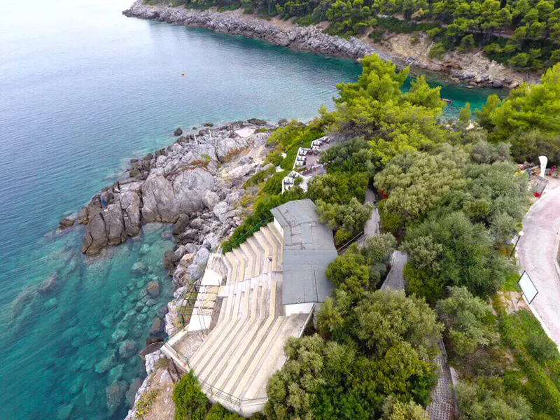Looking down over the stone steps of the amphitheatre to boulders and clear turquoise shallows, olive trees and a path on the slope above
