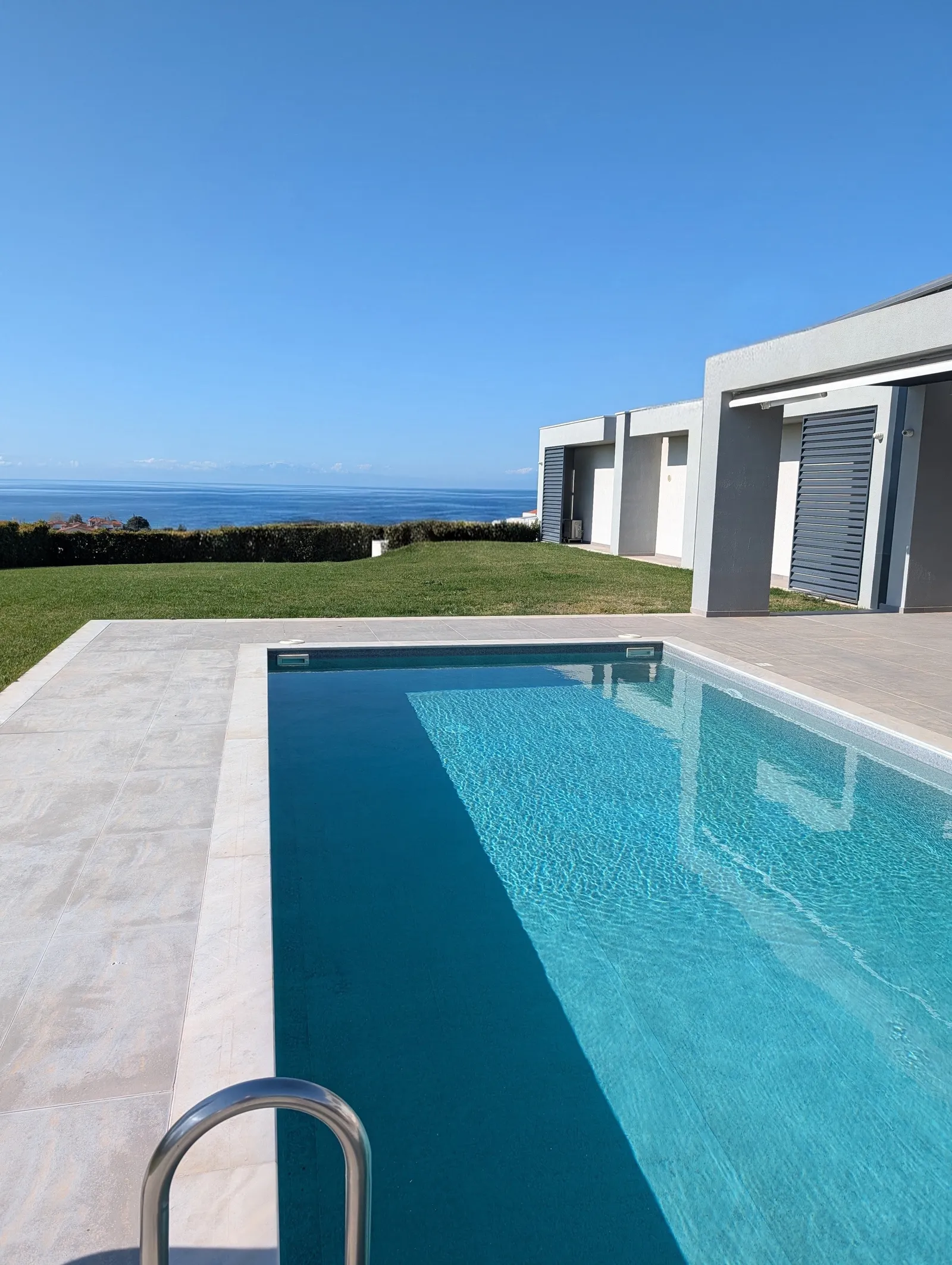A long rectangular pool on a pale tiled terrace beside a modern white villa, lawn running down to a hedge with the Aegean and a far coastline beyond