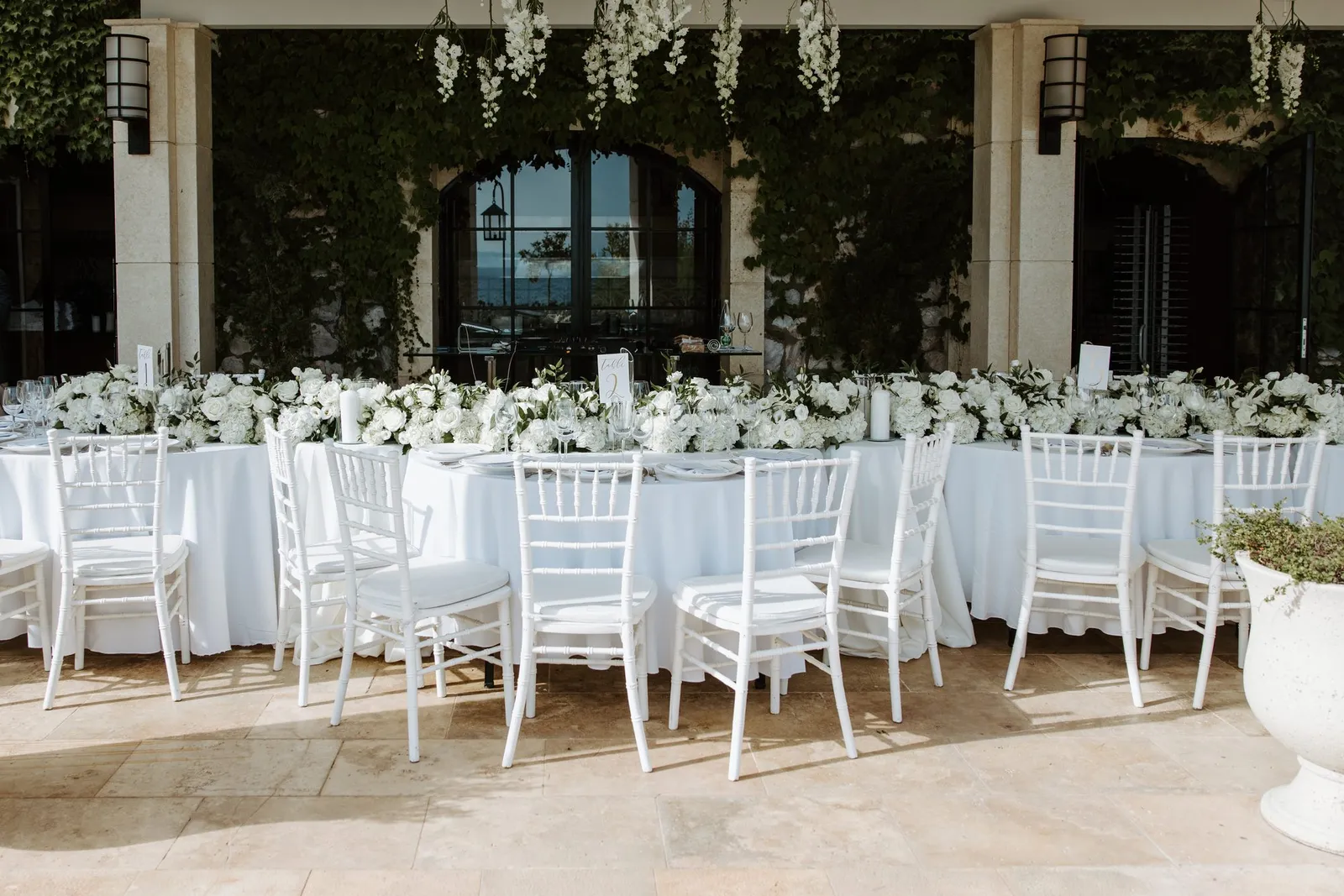 A long table laid beneath white blossom hung from a colonnaded terrace, white chairs down both sides and the sea through the arches beyond