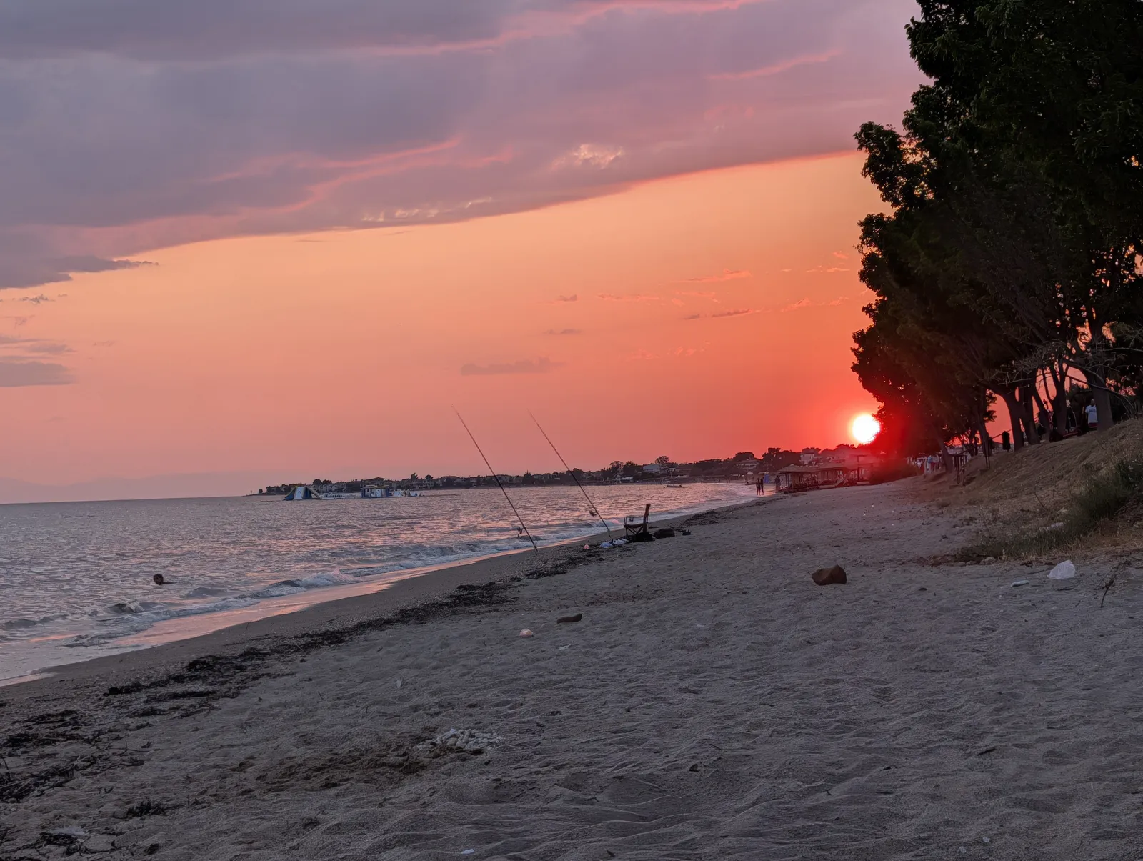 The sun setting behind pines at the edge of a Halkidiki beach, two fishing rods propped at the shoreline